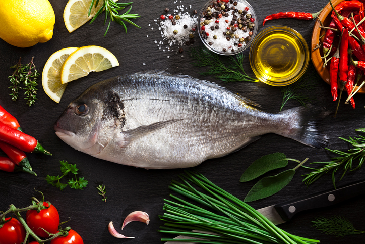 Top view of a raw sea bream fish at the center of an horizontal frame with cooking and seasoning ingredients like olive oil, salt and pepper, rosemary, lemon, thyme, chili pepper, tomatoes, parsley, garlic, chive, are all around the fish. DSRL studio photo taken with Canon EOS 5D Mk II and Canon EF 100mm f/2.8L Macro IS USM